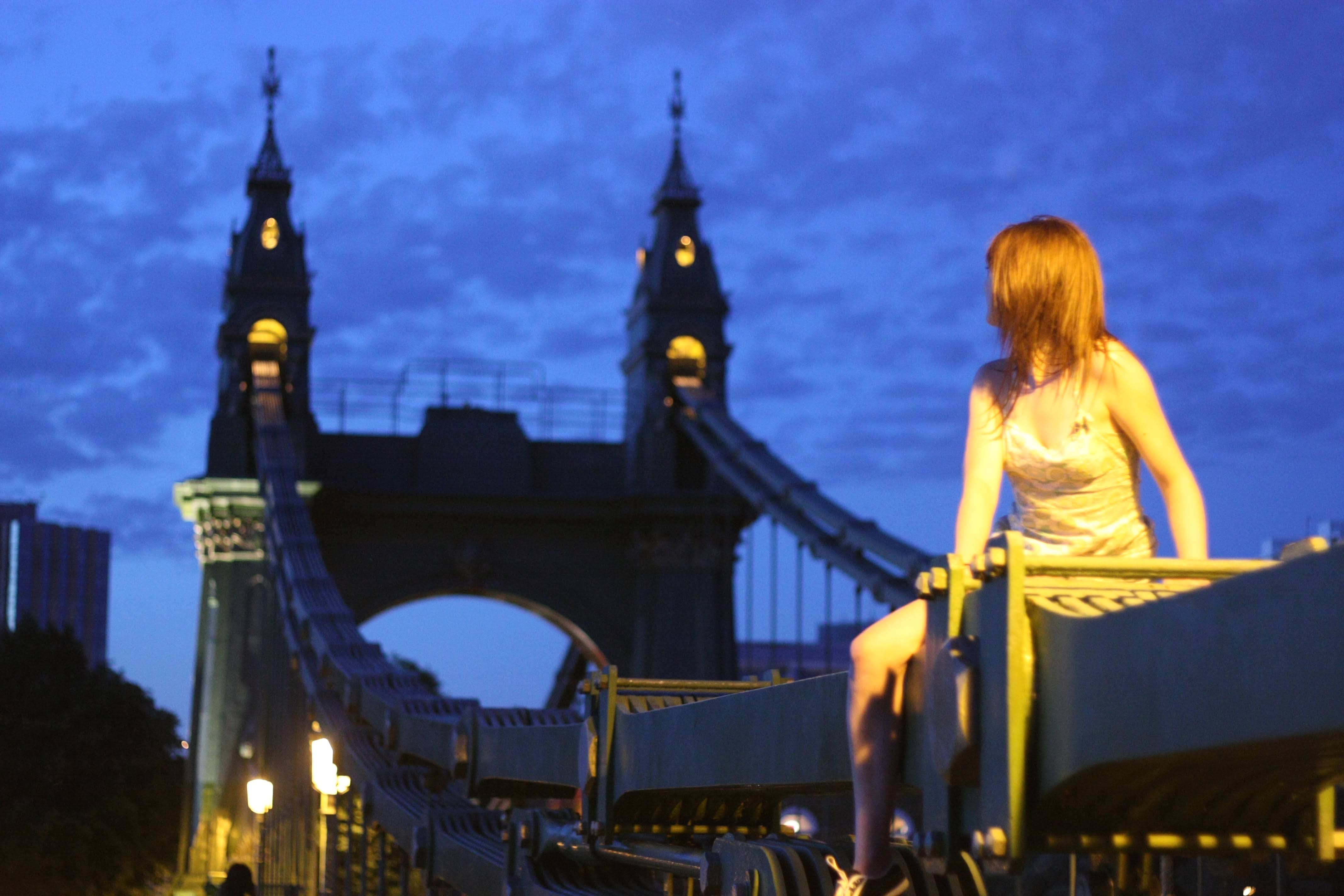 a woman sitting on a bench in front of a bridge ao ~